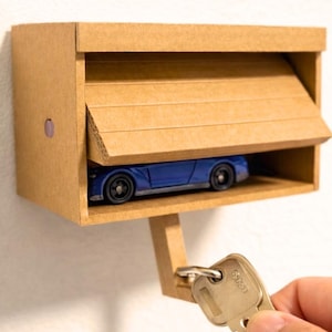 May include: A tan cardboard wall-mounted key holder shaped like a garage with a blue toy car parked inside. The structure features a hinged door panel and a small hook extending from the bottom, which holds a single silver metal key.