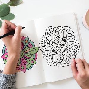 Puede incluir: Una persona coloreando un libro para colorear de mandalas con un marcador negro. El mandala es un diseño circular con intrincados detalles. El libro está abierto en una página con un mandala colorido a la izquierda y un mandala en blanco y negro a la derecha.
