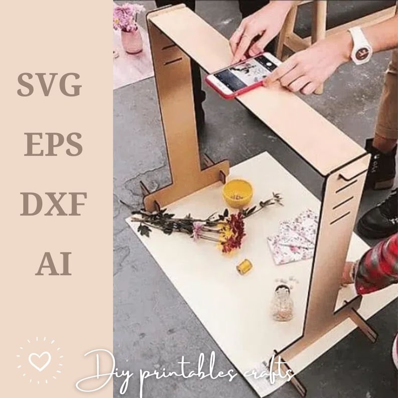 May include: A wooden phone stand with a white background. The stand is made of two vertical pieces of wood connected by a horizontal piece of wood. The stand is designed to hold a phone in a vertical position. The stand is being used to photograph a small bouquet of flowers and other decorative items.