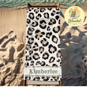 May include: A rectangular beach towel featuring a white background with a repeating black and tan leopard print pattern. The lower section includes a horizontal grey band containing the name Kimberlee printed in a dark grey serif font.