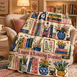 May include: A cream-colored throw blanket featuring a repeating pattern of stacked books, potted flowers, and vases. The text "Laura's Reading Blanket" is printed in the center. The design includes various floral arrangements and colorful book spines.