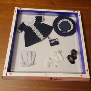 May include: A navy blue velvet doll outfit set in an Effanbee Design Studio box. The set includes a dress with white lace trim and pearl accents, a matching beret, a small handbag, white ruffled bloomers, white socks, and black shoes.
