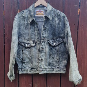 May include: A long-sleeve acid wash denim jacket in a dark gray and light gray mottled pattern. The jacket features a pointed collar, a front button closure, two chest pockets with buttoned flaps, and a label at the neck reading Code Bleu.