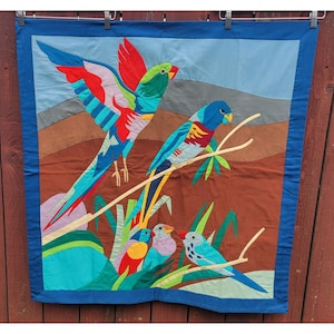 May include: A fabric wall hanging featuring a colorful appliqué scene of five birds. One large bird with red, green, and blue wings is in flight above a perched blue bird. Three smaller birds in red, purple, and blue sit among green foliage.