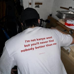 以下が含まれることがあります： 背面に赤い文字で「i'm not kanye west but you'll never find nobody better than me」とプリントされた白い半袖Tシャツ。メインメッセージの下には、同じ赤色のフォントで「ValenVeyra」という小さな文字がプリントされています。