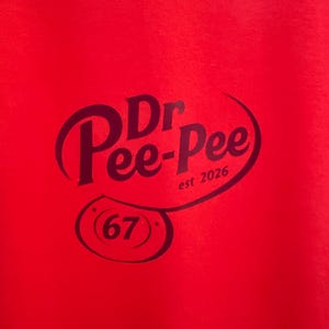 May include: A bright red fabric garment featuring a dark purple graphic print. The design displays the text Dr. Pee-Pee, the phrase est 2026, and a circular emblem containing the number 67, all enclosed within a curved, stylized line.