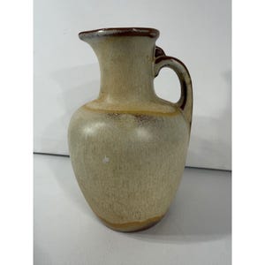 May include: A ceramic pitcher with a rounded body and a flared rim. The surface features a mottled beige and cream glaze with darker brown accents around the rim, the handle, and the base, creating a textured appearance throughout the piece.