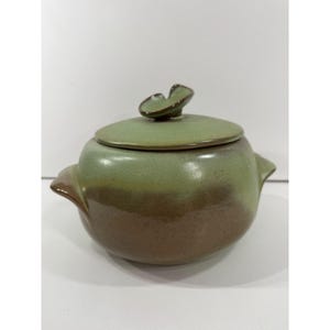 May include: A round ceramic lidded pot featuring a two-tone glaze with a sage green upper section and a brown lower section. The lid includes a small, leaf-shaped handle at the top, and the pot has two small side handles for gripping.