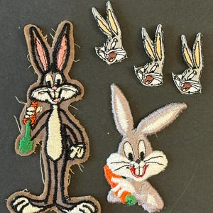 May include: A collection of embroidered fabric patches featuring the cartoon character Bugs Bunny. One large full-body patch shows the character holding a carrot, one medium patch shows the head and shoulders with a carrot, and three small head patches.