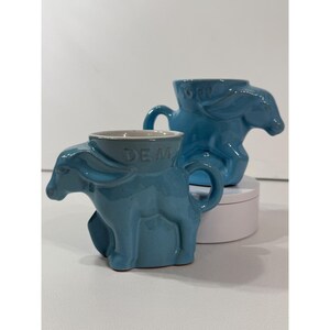 May include: Two light blue ceramic mugs shaped like donkeys. The front mug features the letters DEM embossed on the side, while the rear mug displays the year 1982 embossed on its side. Both mugs have a glossy finish and a sculpted animal design.