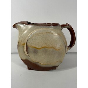 May include: A ceramic pitcher featuring a rounded, circular body with a wide spout and a curved handle. The surface displays a mottled glaze in shades of cream, beige, and dark brown, with horizontal bands of color creating a layered, textured appearance.