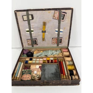 May include: A vintage sewing kit in a rectangular box with a hinged lid. The interior features elastic straps holding needle packets and thread spools. The base contains compartments filled with assorted thread spools, yarn, buttons, and a gray velvet pincushion.