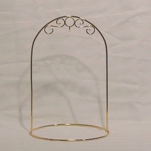 Puede incluir: Un expositor de metal dorado con base circular y parte superior arqueada. La parte superior presenta adornos decorativos. El soporte está diseñado para exhibir objetos.