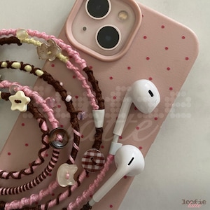May include: A pink smartphone case with red polka dots featuring a coiled cord accessory. The cord is wrapped in pink and brown thread, decorated with small beads, a cream star-shaped button, and a brown plaid button, attached to white earbuds.