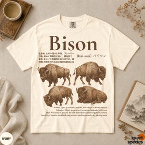 Può includere: Una t-shirt a maniche corte color avorio con una grande grafica marrone raffigurante quattro illustrazioni di bisonti. Il testo include la parola Bison, la pronuncia fonetica e dettagliati fatti biologici in inglese e giapponese stampati in marrone.