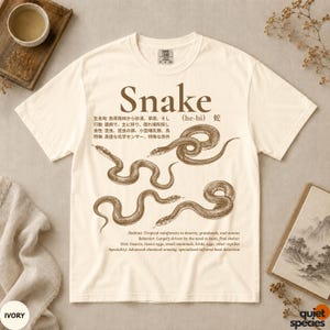 Puede incluir: Una camiseta de manga corta color marfil con un estampado gráfico marrón de tres serpientes. El texto incluye la palabra Snake, caracteres japoneses y descripciones en inglés sobre el hábitat, comportamiento y dieta.