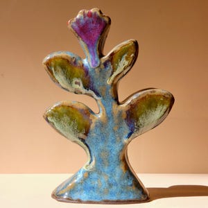 May include: A ceramic sculpture shaped like a stylized plant with a central stem and four symmetrical leaves. The piece features a glossy glaze in shades of deep blue, olive green, and brown, with a vibrant purple accent on the top flower.