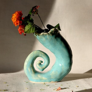 May include: A turquoise ceramic vase with a spiraled, horn-like shape and a ruffled opening. The vase features a glossy, uneven glaze and contains a small cluster of vibrant orange flowers with green leaves extending from the top opening.
