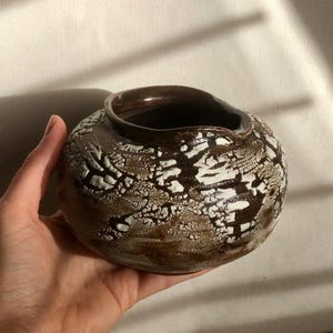 May include: A small, round ceramic bowl with a wide opening and a short, curved spout. The exterior features a textured, crackled glaze in shades of dark brown and cream, creating a high-contrast, web-like pattern across the entire surface.