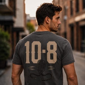 May include: A charcoal gray short-sleeve t-shirt featuring a large tan graphic on the back. The design displays the text 10-8 in a bold athletic font above a detailed illustration of a vintage-style handheld radio microphone with a coiled cord.