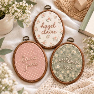 Könnte beinhalten: Drei ovale Holzstickrahmen mit Stoff, auf dem Namen in Schreibschrift gestickt sind. Ein Rahmen hat cremefarbenen Blumenstoff mit hazel claire, einer hat rosa gepunkteten Stoff mit luna june und einer hat grünen Gänseblümchenstoff mit willow rae.