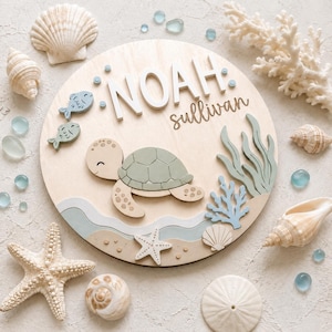 Può includere: Una targa da parete circolare in legno con il nome NOAH Sullivan in lettere bianche e corsive. Il design include una tartaruga marina verde chiaro, due piccoli pesci blu, corallo azzurro e strati ondulati.