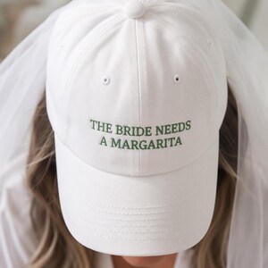 Może przedstawiać: Biała czapka z daszkiem z wyhaftowanym zielonym napisem THE BRIDE NEEDS A MARGARITA na przednim panelu. Czapka ma usztywnianą koronę, zakrzywiony daszek, guzik na górze oraz przymocowany z tyłu przezroczysty biały welon.