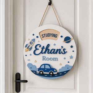 May include: A circular white wooden door sign featuring the text Ethan's Room and Studying. The design includes a blue car on a road, a rocket ship, a planet with rings, and various stars in shades of blue and yellow, all suspended by a tan rope.