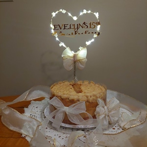 May include: A round cake topped with a heart-shaped wire frame illuminated by small white lights. The frame features a gold-coloured cutout reading "EVELYN'S 15th Birthday" and a sheer white ribbon bow. The cake is decorated with a white ribbon.