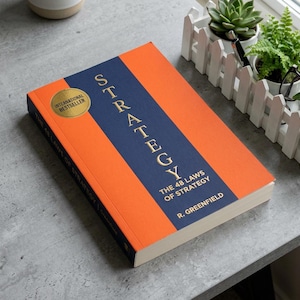 May include: A hardback book titled Strategy: The 48 Laws of Strategy by R. Greenfield. The cover features a bold orange and dark blue vertical design with gold lettering and a circular gold International Bestseller seal on the front cover.