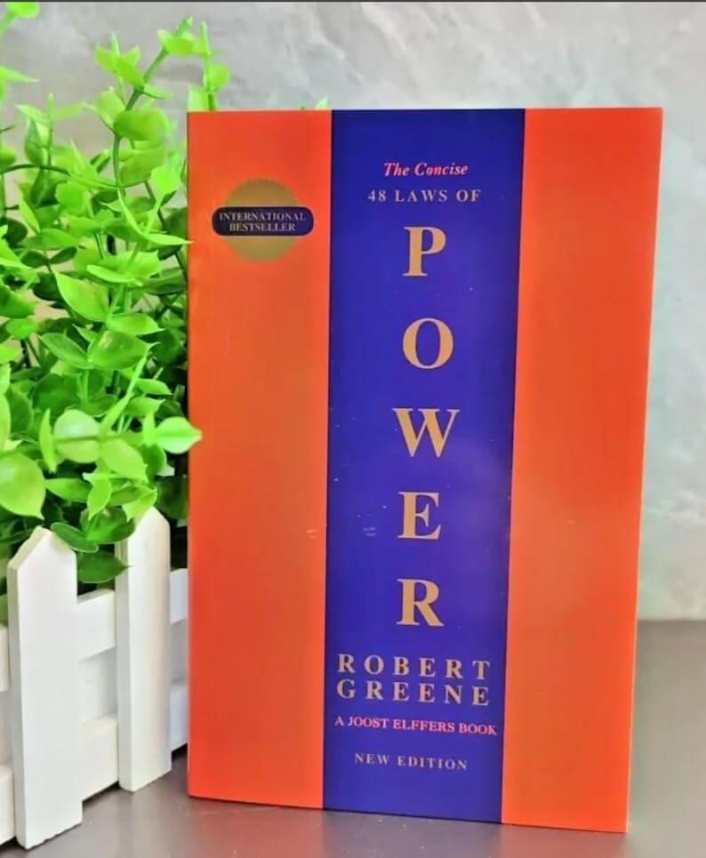 May include: A book titled The Concise 48 Laws of Power by Robert Greene, featuring a vertical blue central band on an orange cover. The text includes International Bestseller, A Joost Elffers Book, and New Edition in white and gold lettering.