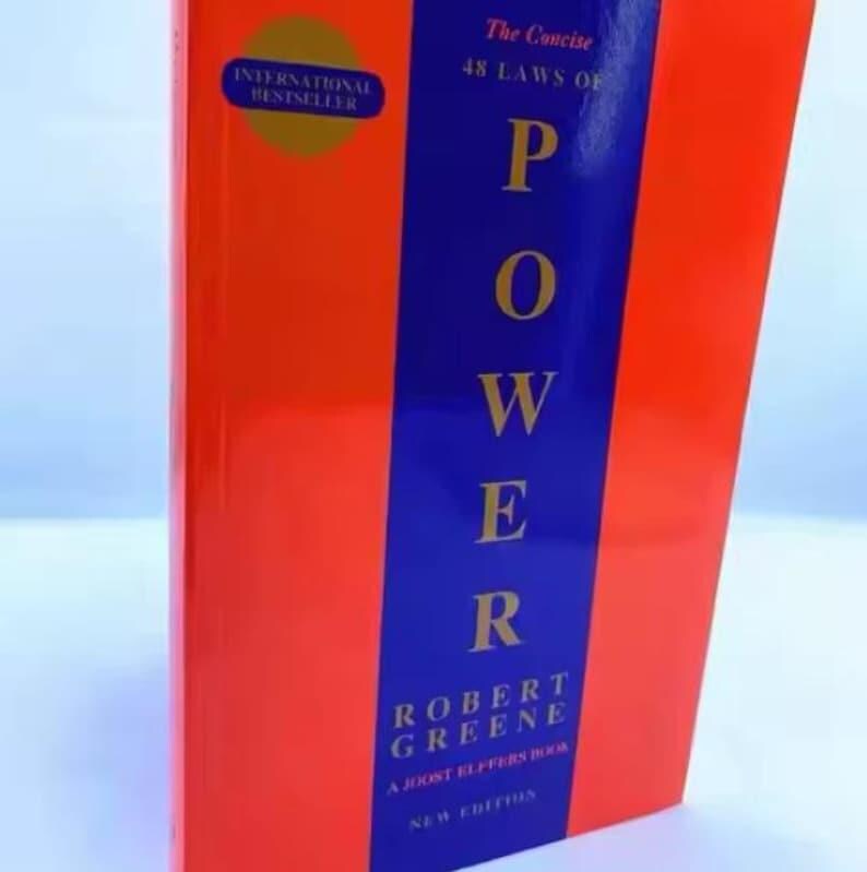 May include: A book titled The Concise 48 Laws of Power by Robert Greene and Joost Elffers. The cover features a bright orange background with a central vertical blue stripe. The title is printed in gold lettering, and a gold circle badge reads International Bestseller.