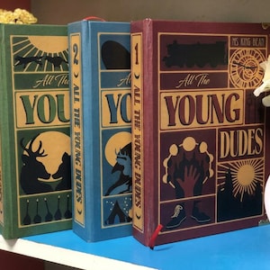 Może przedstawiać: Trzy książki w twardej oprawie zatytułowane All The Young Dudes autorstwa Ms King Bean, ponumerowane od jednego do trzech na grzbietach. Okładki zawierają graficzne ilustracje w kolorze zielonym, niebieskim i bordowym z sylwetkami zwierząt, pociągu i zegara.