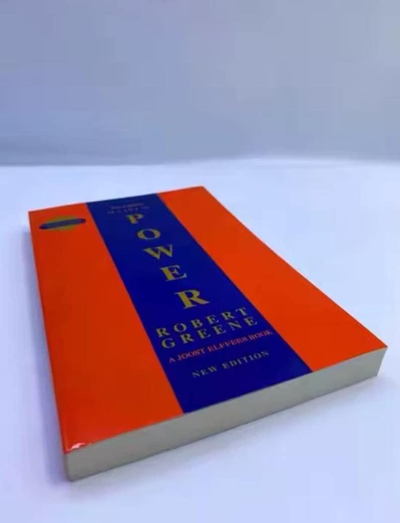 May include: A paperback book titled The 48 Laws of Power by Robert Greene, a Joost Elffers book, new edition. The cover features a wide vertical blue stripe centred on an orange background with yellow text for the title and author name.