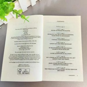 May include: An open book displaying the contents page and copyright information. The right page lists laws from 1 to 9, including titles like Never Outshine the Master. The left page contains publication details, ISBN numbers, and a FSC certification logo.