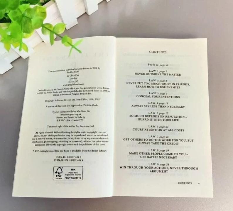 May include: An open book displaying the contents page and copyright information. The right page lists laws from 1 to 9, including titles like Never Outshine the Master. The left page contains publication details, ISBN numbers, and a FSC certification logo.