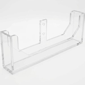 Puede incluir: Un estante de exhibición de pared hecho de acrílico transparente con una base rectangular, dos paneles laterales y una pestaña de soporte vertical central con dos orificios de montaje circulares. La estructura es transparente y rígida.