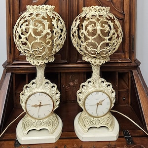 May include: A pair of cream-colored table lamps with integrated clock faces at the base. Each lamp features a spherical, open-work filigree shade and a base with Roman numerals. They are positioned in front of a tall, dark wood cabinet with floral art.