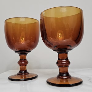 May include: Two amber-colored glass goblets with a smooth, rounded bowl and a thick, multi-tiered stem leading to a circular base. The glass is translucent and features a warm, golden-brown hue, with one goblet slightly larger than the other.