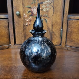 May include: A spherical black glass perfume bottle with a tall, pointed stopper. The glass features a mottled, metallic gold and bronze speckled pattern across its rounded body, creating a reflective surface that contrasts with the solid black neck.