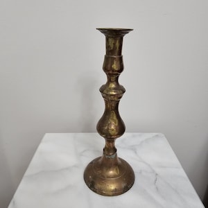 May include: A tall, tarnished brass candlestick holder featuring a multi-tiered, turned stem design with a wide, circular base and a flared top rim. The metal surface displays a weathered, aged patina with visible dark spots and metallic texture.