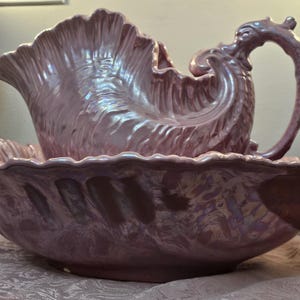 May include: A lavender-colored ceramic wash basin and matching pitcher set with an iridescent glaze. The pitcher features a shell-like, fluted body and a curved handle, while the basin has a wide, scalloped rim and a textured, wavy surface.
