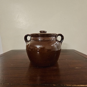 May include: A dark brown glazed ceramic bean pot with a matching lid and two side handles. The pot features a rounded body and a smooth, reflective surface. The lid has a small central knob for lifting, and the handles are curved and sturdy.