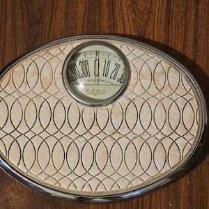 May include: An oval-shaped Borg bathroom scale with a cream-colored surface featuring a repeating black interlocking oval pattern. The scale has a circular analog dial with black numbers and the text BORG, THE BORG-ERICKSON CORP, CHICAGO, U.S.A.