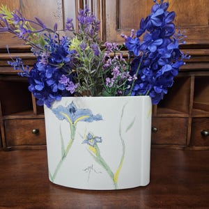 May include: A white ceramic vase with a flat, semi-circular profile, decorated with hand-painted blue and yellow iris flowers and green stems. The vase is filled with a vibrant arrangement of artificial blue and purple flowers.