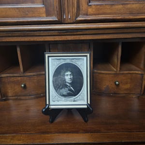 May include: A framed black and white portrait print featuring a person with long hair in an oval border. The rectangular frame has a thin gold inner edge and a black outer border, resting on a black plastic display stand for upright viewing.