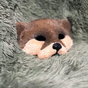 May include: A plush animal mask featuring a brown upper head and ears with a light peach-colored muzzle area. The mask has two dark, oval-shaped eye openings and a small, shiny black nose centered on the lower portion of the face.