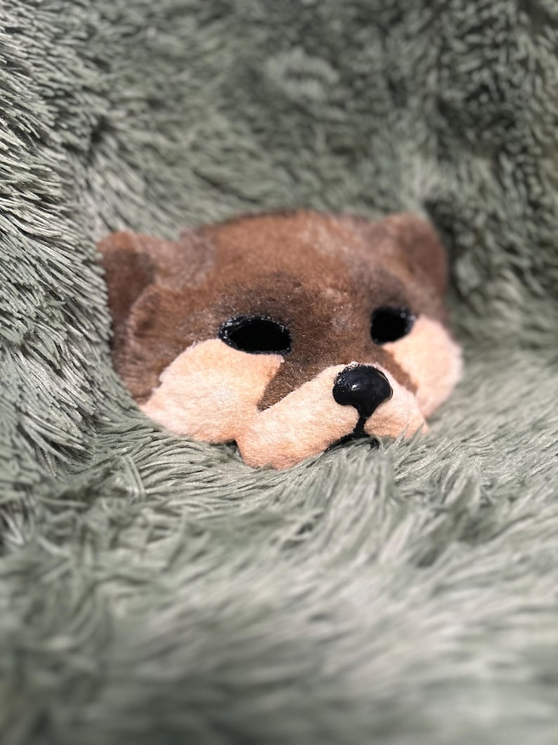 May include: A plush animal mask featuring a brown upper head and ears with a light peach-colored muzzle area. The mask has two dark, oval-shaped eye openings and a small, shiny black nose centered on the lower portion of the face.