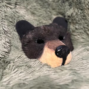 May include: A black bear mask featuring dark fur, rounded ears, and a tan muzzle. The mask has two eye openings and a prominent, glossy black nose with a vertical line extending down the center of the tan snout area, creating a realistic look.