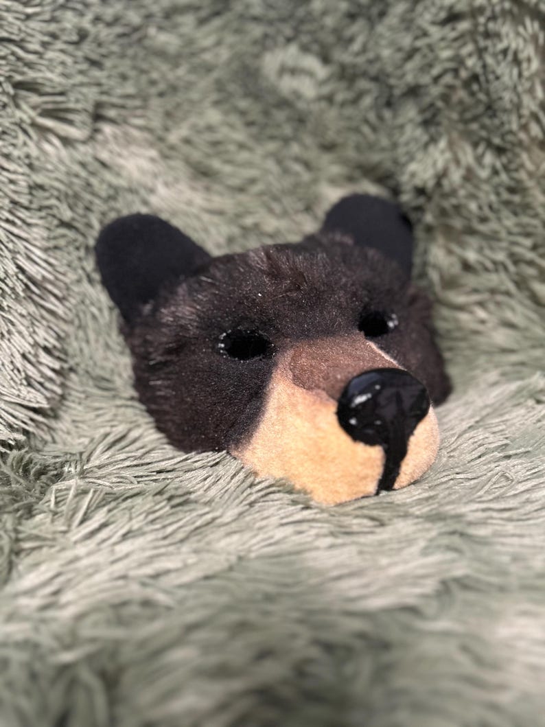 May include: A black bear mask featuring dark fur, rounded ears, and a tan muzzle. The mask has two eye openings and a prominent, glossy black nose with a vertical line extending down the center of the tan snout area, creating a realistic look.